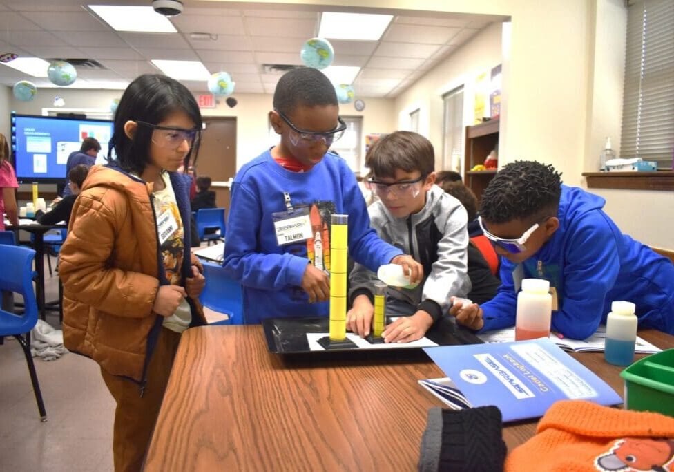 Students gathered around a table measuring liquid in graduated cylinders during a hands-on activity.